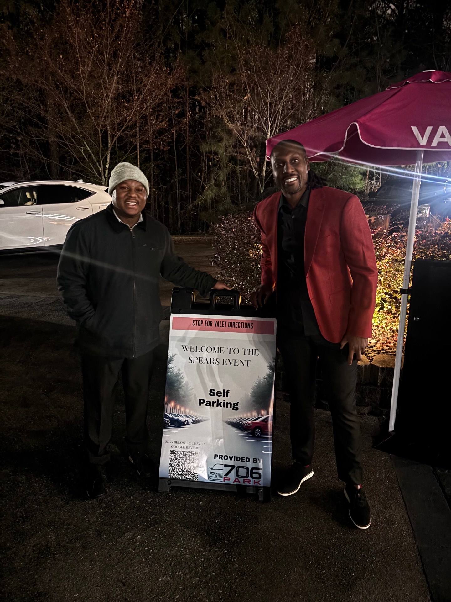 Two 706 Park team members standing with a valet directions sign under a red valet umbrella at a night event