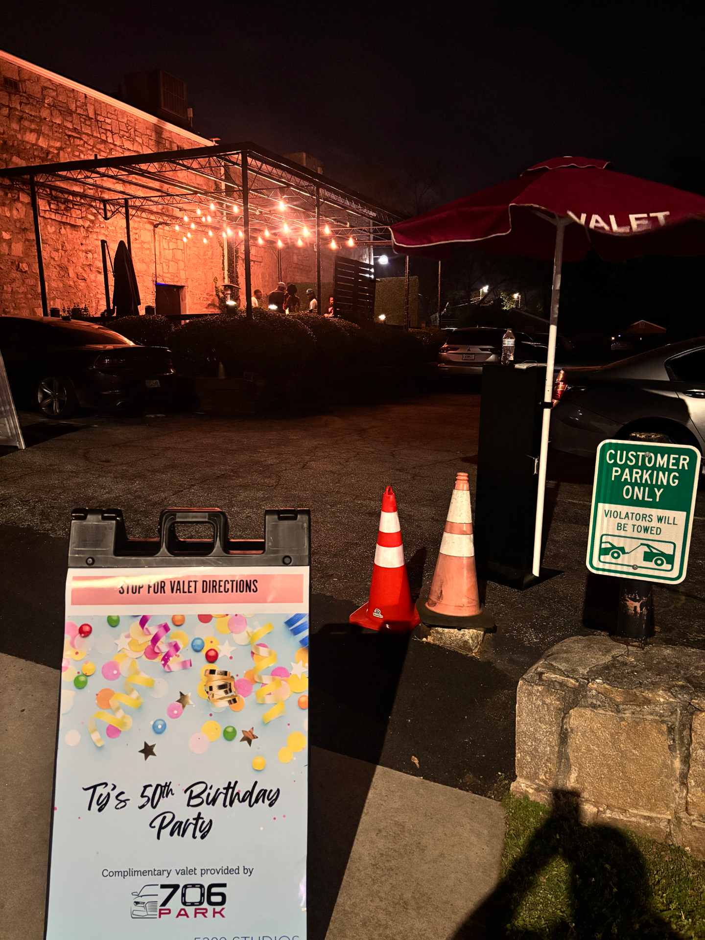 706 Park stop-for-valet-directions sign for a 50th birthday party beside a red VALET umbrella and valet stand in a small lot at night