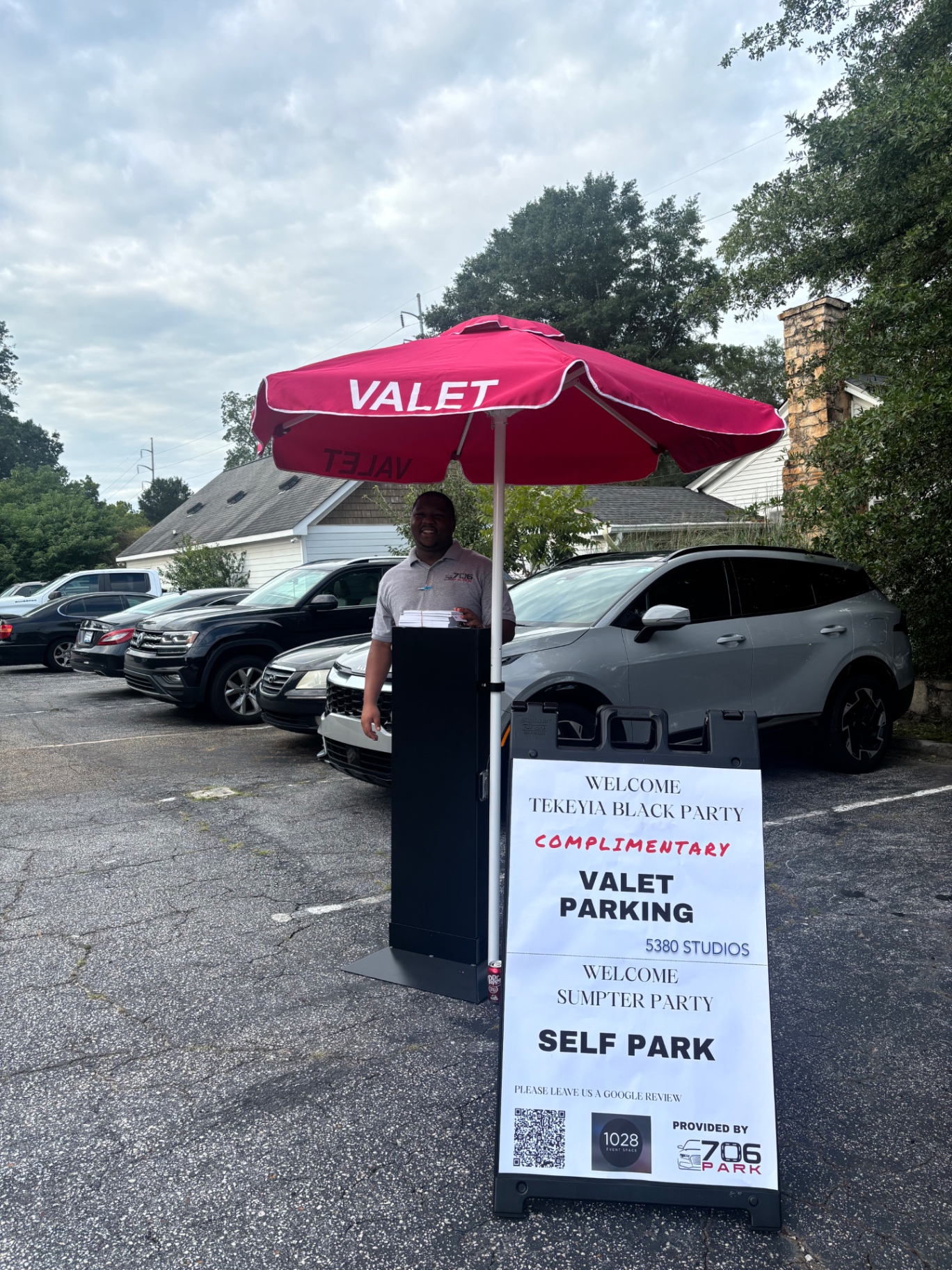 706 Park valet attendant at a stand under a red VALET umbrella in a compact event space parking lot filled with cars